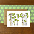 Load image into Gallery viewer, A white greeting card featuring an illustration of birch or aspen trees with green leaves, placed in front of a brown kraft envelope on a green polka-dot sheet, all set on a wooden surface.