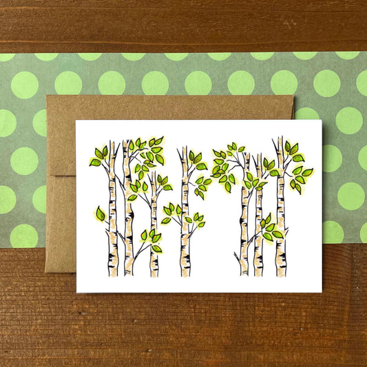 A white greeting card featuring an illustration of birch or aspen trees with green leaves, placed in front of a brown kraft envelope on a green polka-dot sheet, all set on a wooden surface.